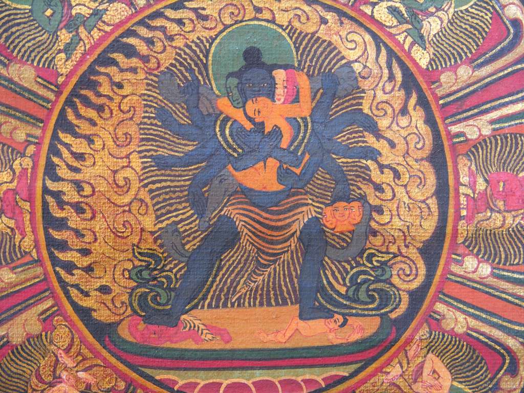 Chakrasamvara Mandala 11 Centre Close Up The centre of the mandala shows the blue-black Chakrasamvara with four faces and twelve arms, the two main arms embracing his consort Vajravarahi, who has one face and two arms. He wears a girdle made of tiger skin and the skin of an elephant, grasping with one of his right hands the left paw while one of his left hands holds the left foot. With his right leg set forward he presses down the head of Kalabhairava, and his left leg presses the breast of Kali. In one of his left hands he holds the four severed heads of god Brahma.